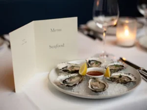 Open à la carte menukaart op wit linnen tafelkleed naast bord met verse oesters en citroen bij kaarslicht in een fine dining restaurant.