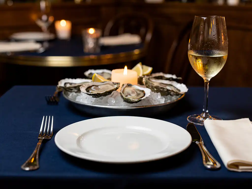 Gedekte tafel in Amsterdams brasserie met verse oesters op crushed ice, zilveren bestek, wit bord en glas witte wijn bij kaarslicht.