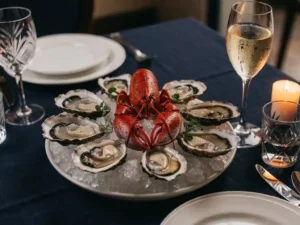 Hele gekookte kreeft omringd door verse oesters op crushed ice, kruidengarnituur en glas witte wijn bij kaarslight op marineblauw linnen.