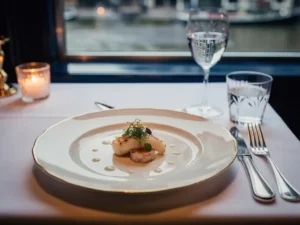 Elegant zeevruchtengerechtje op porseleinen bord met zilver bestek, kaarslicht en Amsterdams grachtenraam op de achtergrond.