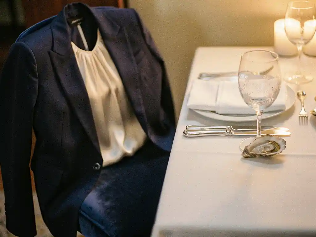 Marine blazer en zijden blouse over fluwelen stoel bij gedekte eettafel met kristallen wijnglas, zilverwerk en kaarsenlicht op wit linnen.