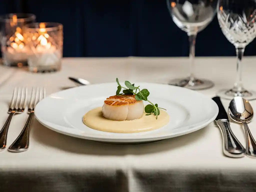Elegant opgemaakte scallop met microgreens en bisque reductie op wit bord, zilveren bestek en kaarslicht op de achtergrond.