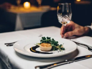 Gebakken sint-jakobsschelp met microgreens en sausreductie op wit bord, geserveerd in een intiem Amsterdams restaurant bij kaarslicht.