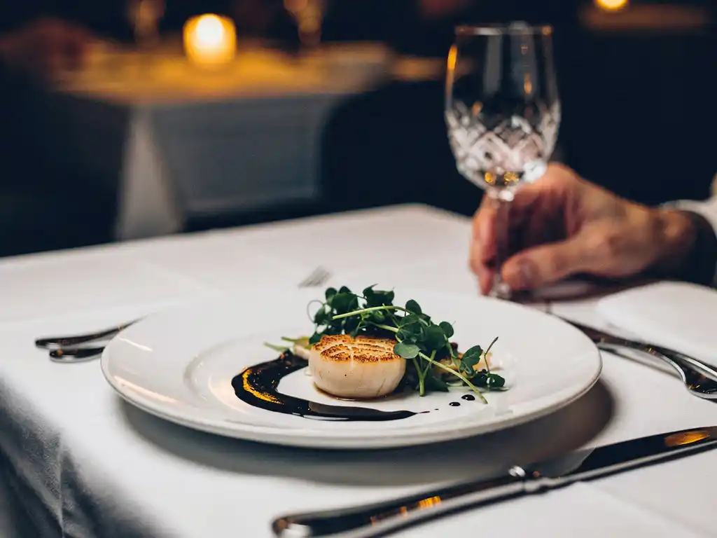 Gebakken sint-jakobsschelp met microgreens en sausreductie op wit bord, geserveerd in een intiem Amsterdams restaurant bij kaarslicht.