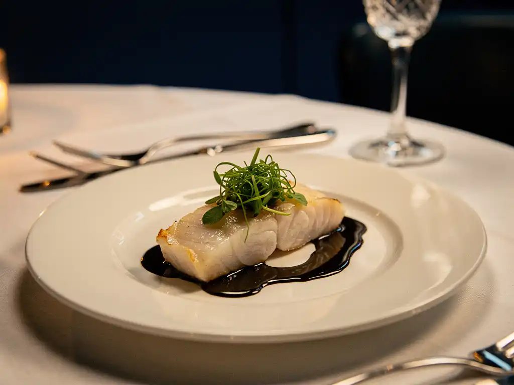 Fijn gerecht met visfilet, microgreens en glanzende saus op wit bord, kaarslicht en kristallen wijnglas op achtergrond.