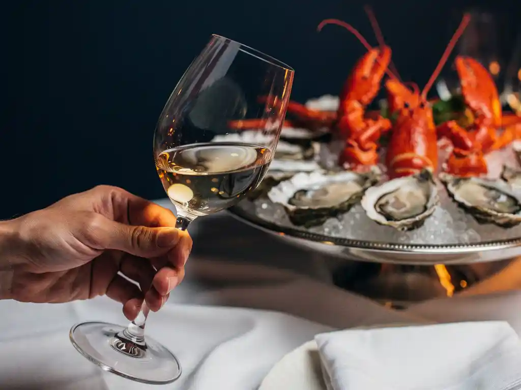 Sommelier kantelt kristallen wijnglas met goudgele witte wijn, op de achtergrond een zeevruchtenplateau met oesters en kreeft bij kaarslicht.
