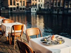 Terras aan Amsterdamse gracht met verse oesters op tafel, houten bistrostoelen en witte tafellakens, historische grachtenpanden weerspiegeld in het water.