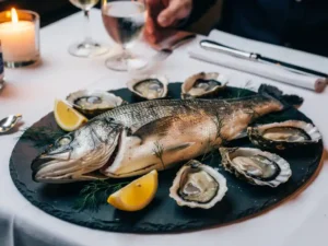Hele zeebaars op leisteen bord met oesters, citroenpartjes en dille in een elegante Amsterdamse brasserie bij kaarslicht.